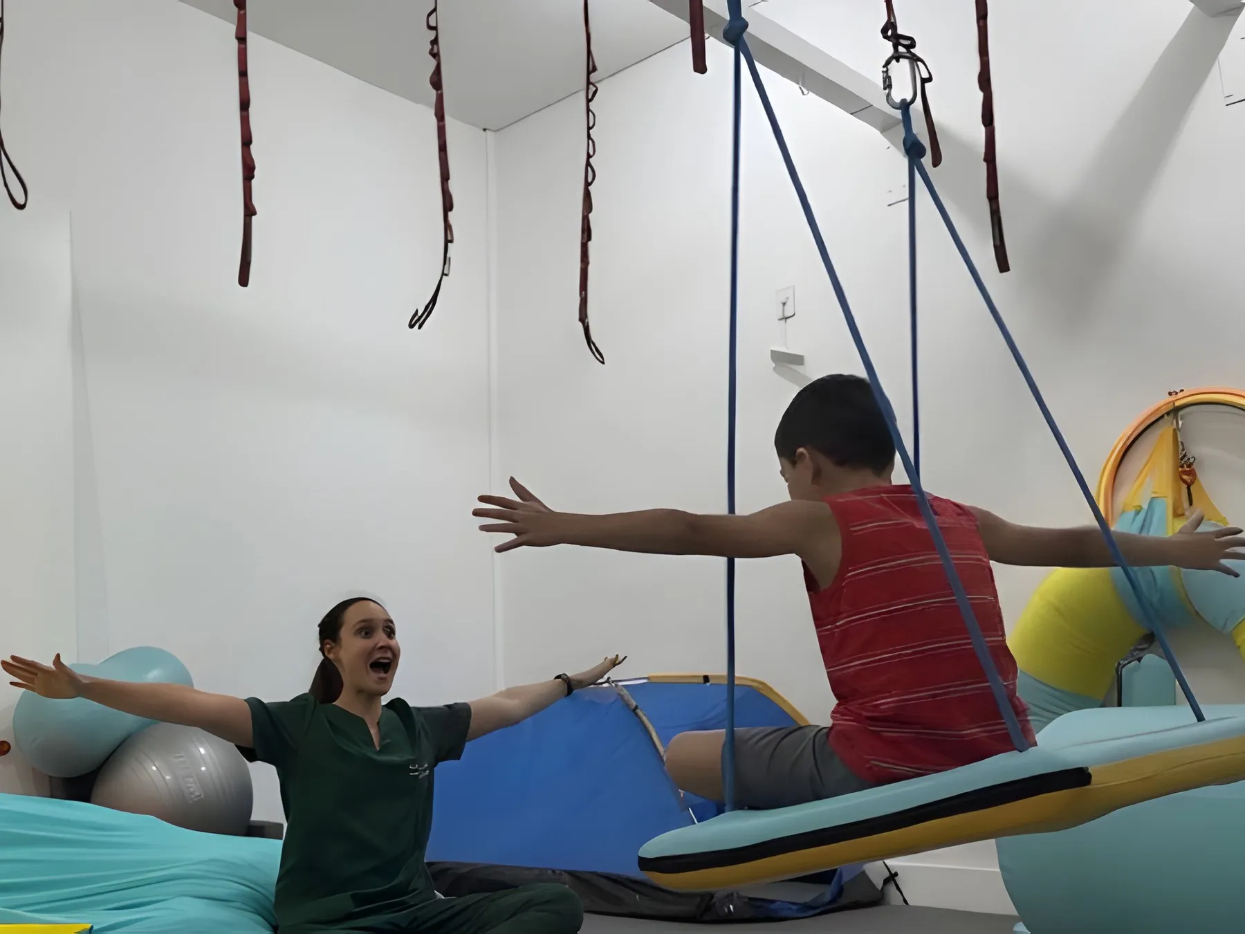 Terapeuta sentada no chão abre os braços para um menino, de costas, que se equilibra de braços abertos no balanço de plataforma