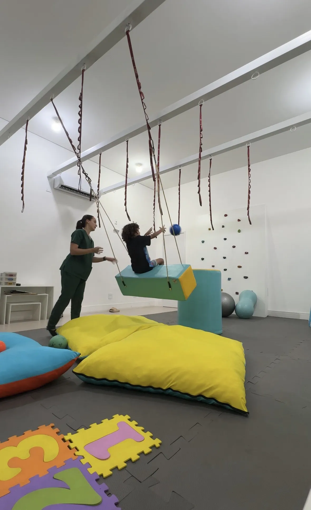 Terapeuta do Espaço Akatu empurrando o balanço suspenso em que uma criança, de costas, brinca de pegar uma bola
