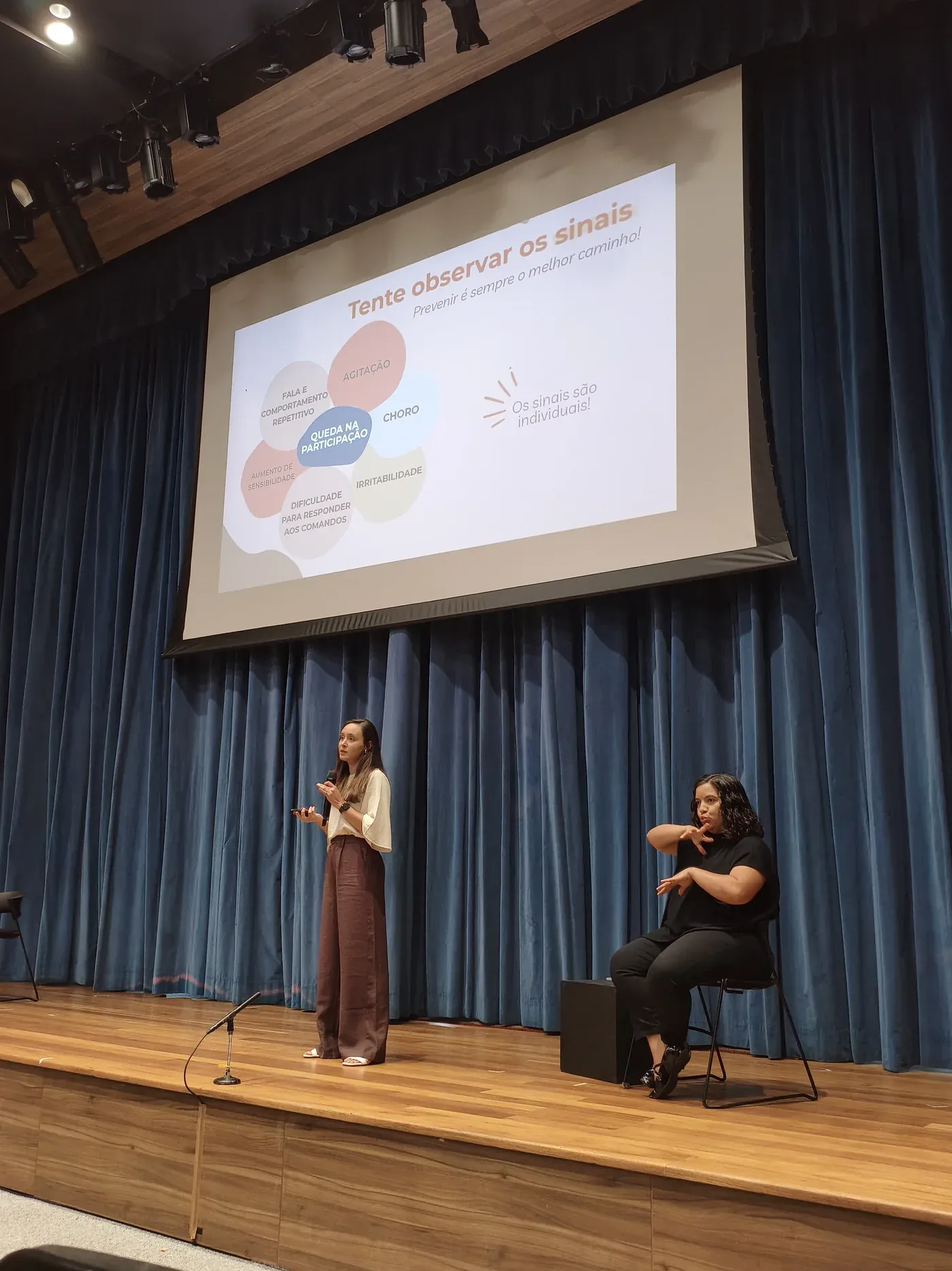 Natália Maia conduzindo o treinamento no auditório, ao lado de intérprete de Libras, com slide sobre observar os sinais