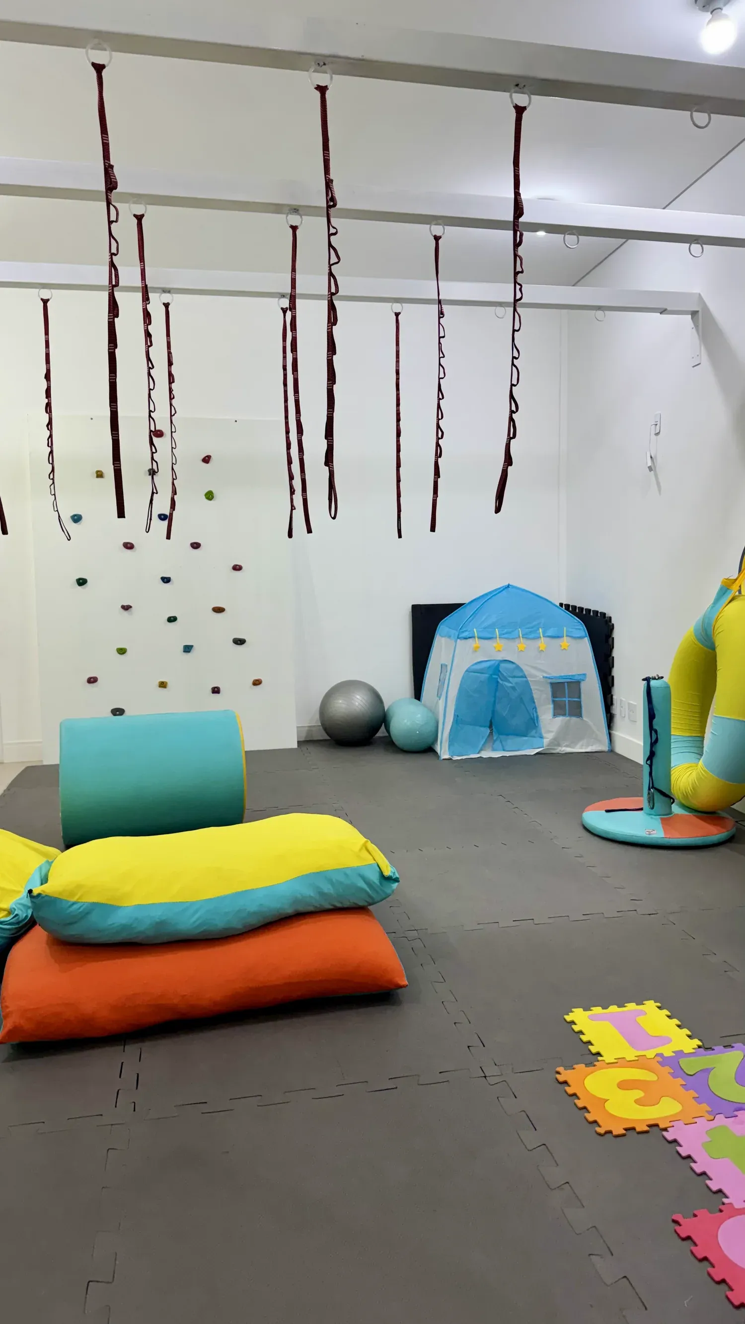 Sala de integração sensorial vazia: parede de escalada, cordas suspensas, tenda e almofadões coloridos