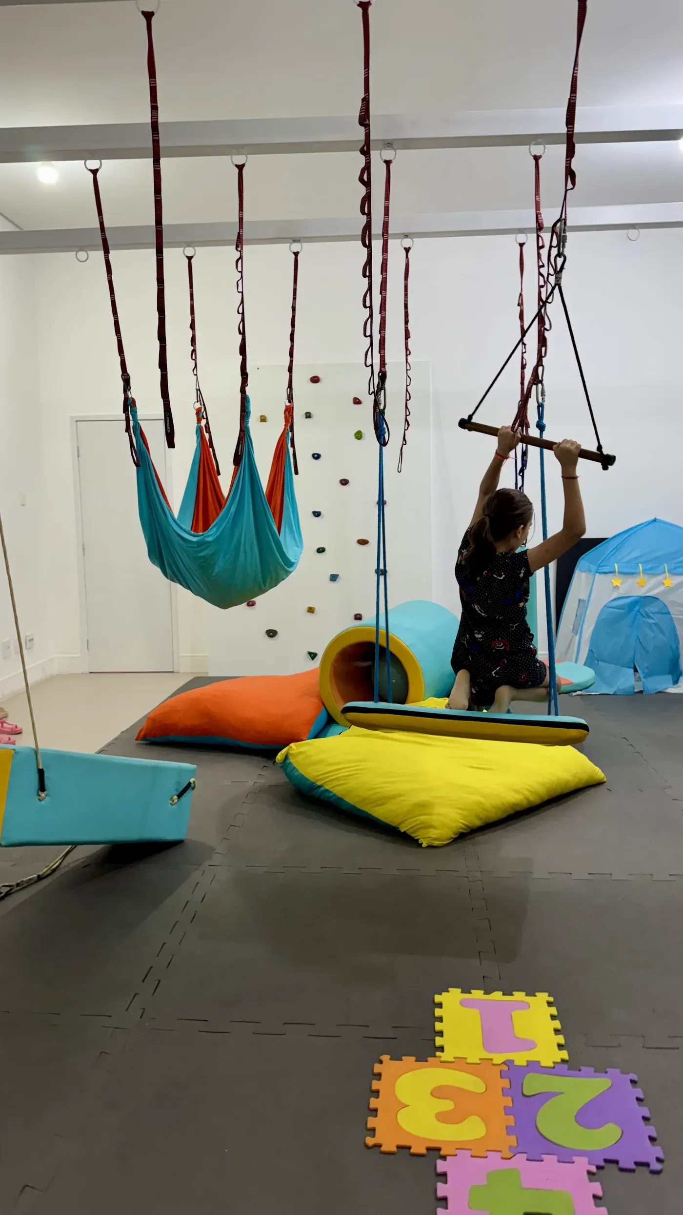 Criança de costas no trapézio da sala de integração sensorial, com rede de tecido suspensa ao lado