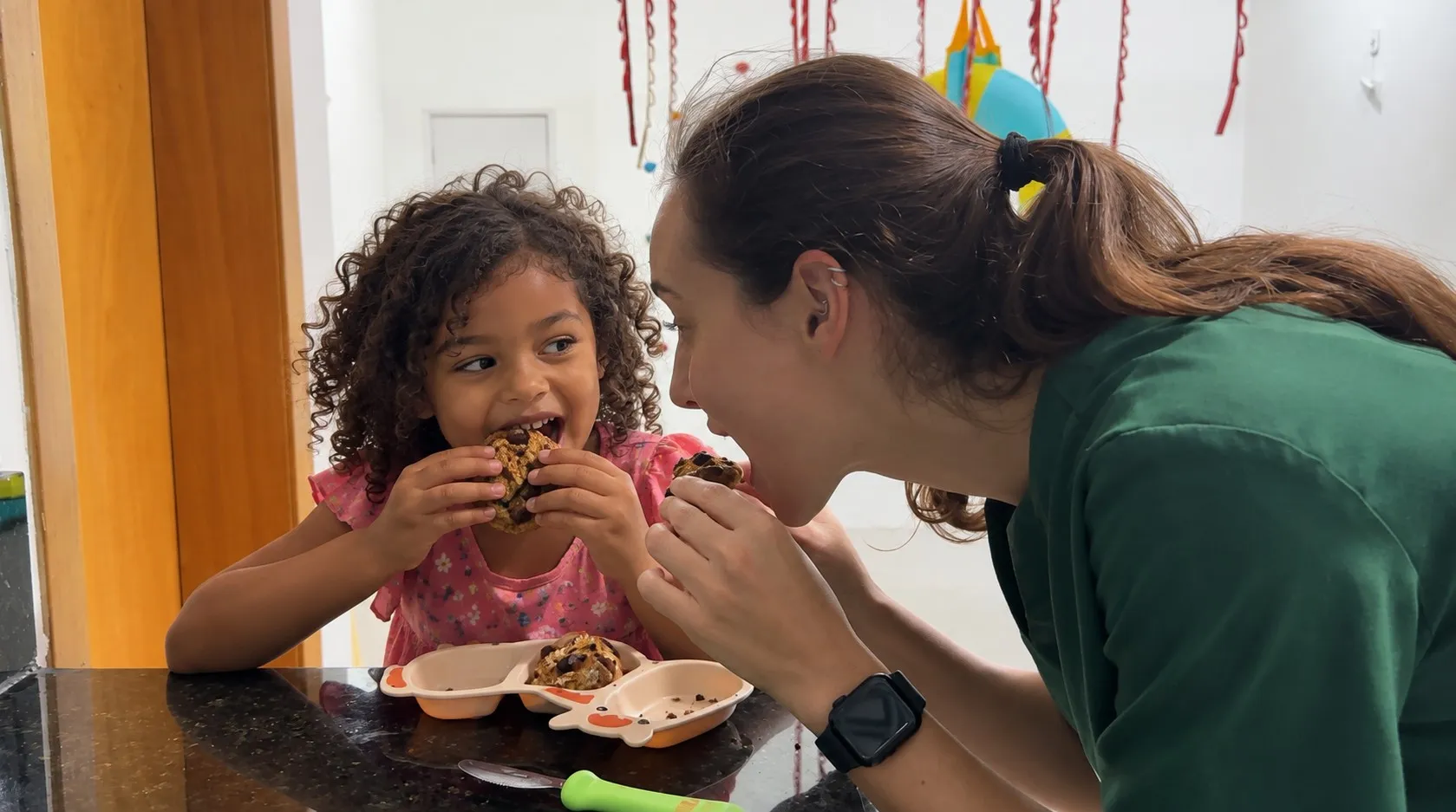 Terapeuta e menina comendo juntas um bolinho à mesa, em sessão de terapia alimentar